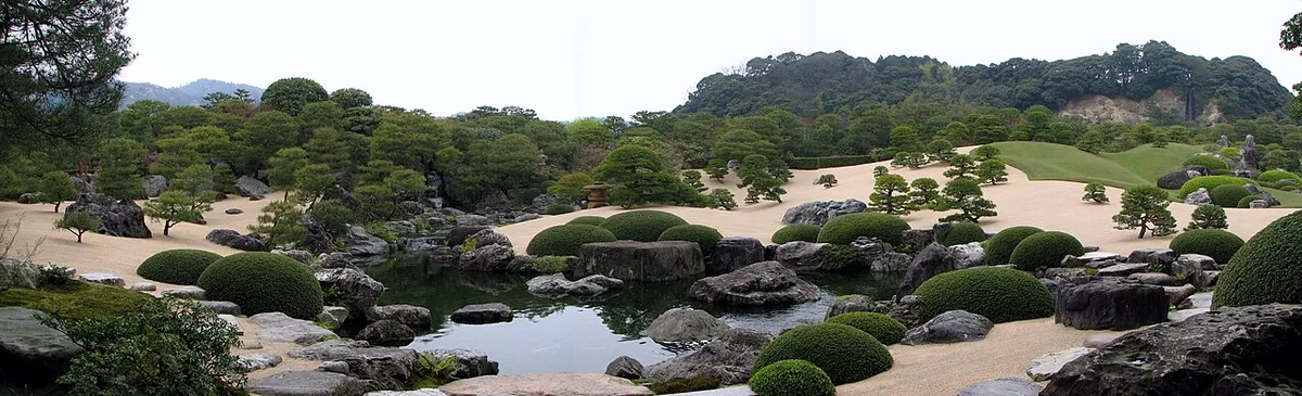 Panorama of a garden pond with rocks and rounded shrubs, white gravel, pines and borrowed wooded hills