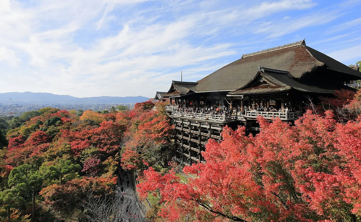 Red and orange maples around the wooden stage of Kiyomizu-dera temple in Kyoto, with visitors on the stage and the city behind