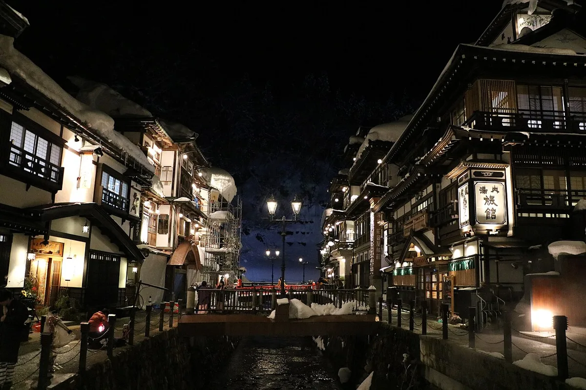 Ginzan Onsen at night in winter: snow on the roofs of multi-storey wooden ryokan on both banks of the river, lit by street lamps