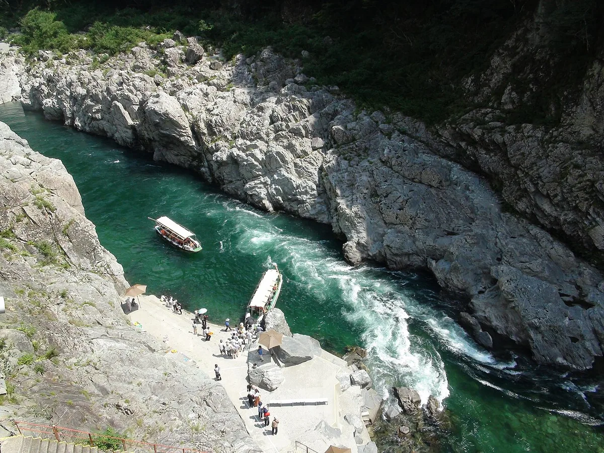 Two sightseeing boats on the emerald Yoshino River between grey rock walls in Oboke Gorge, with passengers waiting on the landing