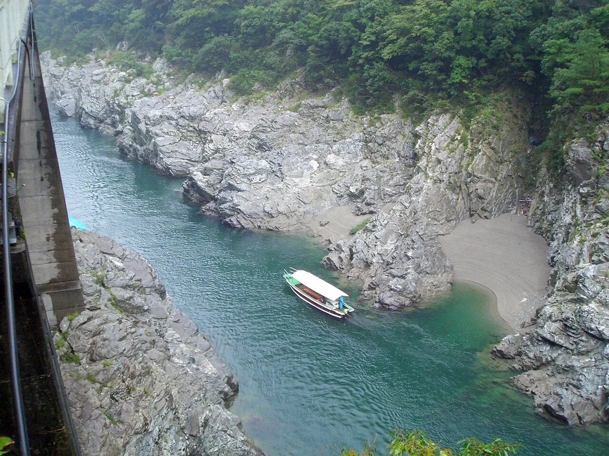 Green river between grey rock walls with a covered sightseeing boat, seen from a roadside railing above the gorge