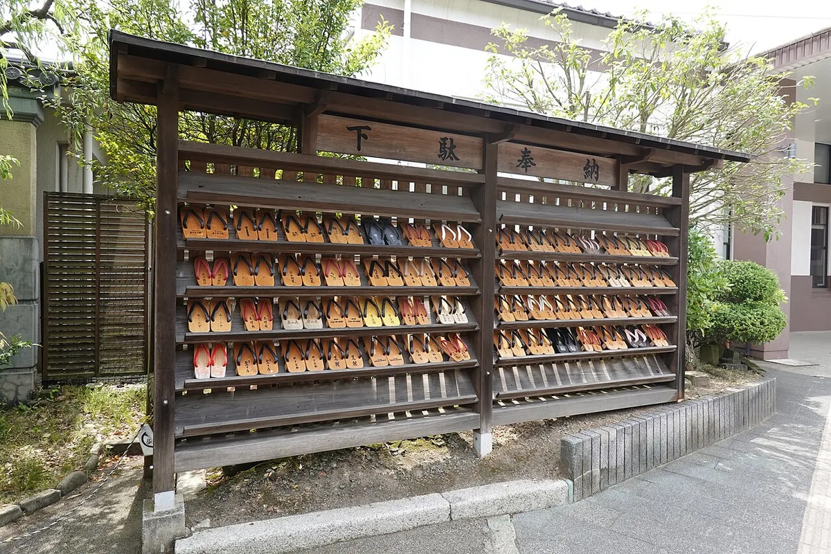 Wooden rack of geta sandals with a sign reading geta offering, beside a paved street