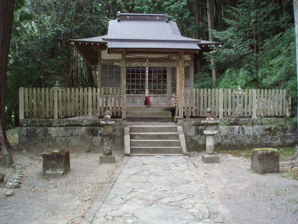 Small wooden shrine hall behind a low fence and stone lanterns in the forest at Takijiri-oji