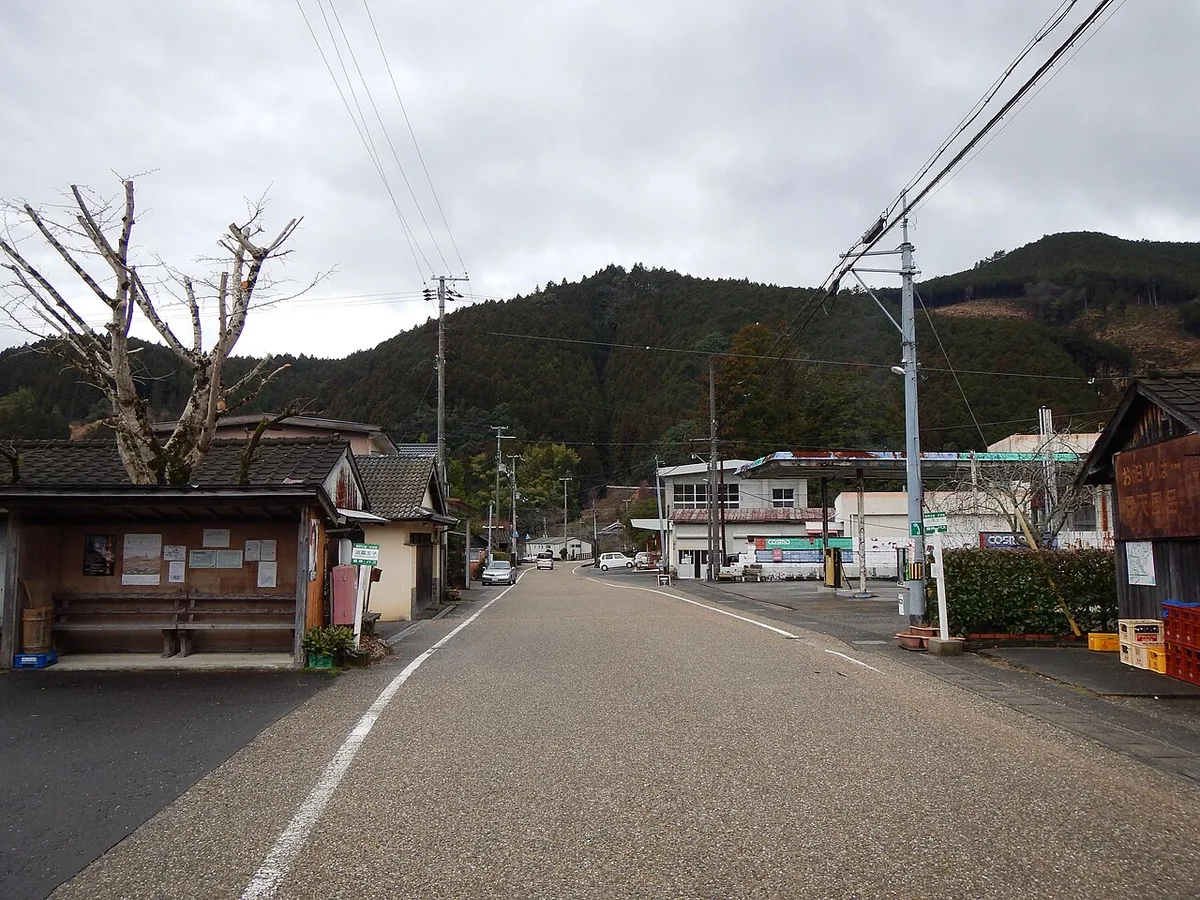 A quiet village road at Gyuba-doji-guchi with a wooden shelter, bare trees and forested hills under grey sky