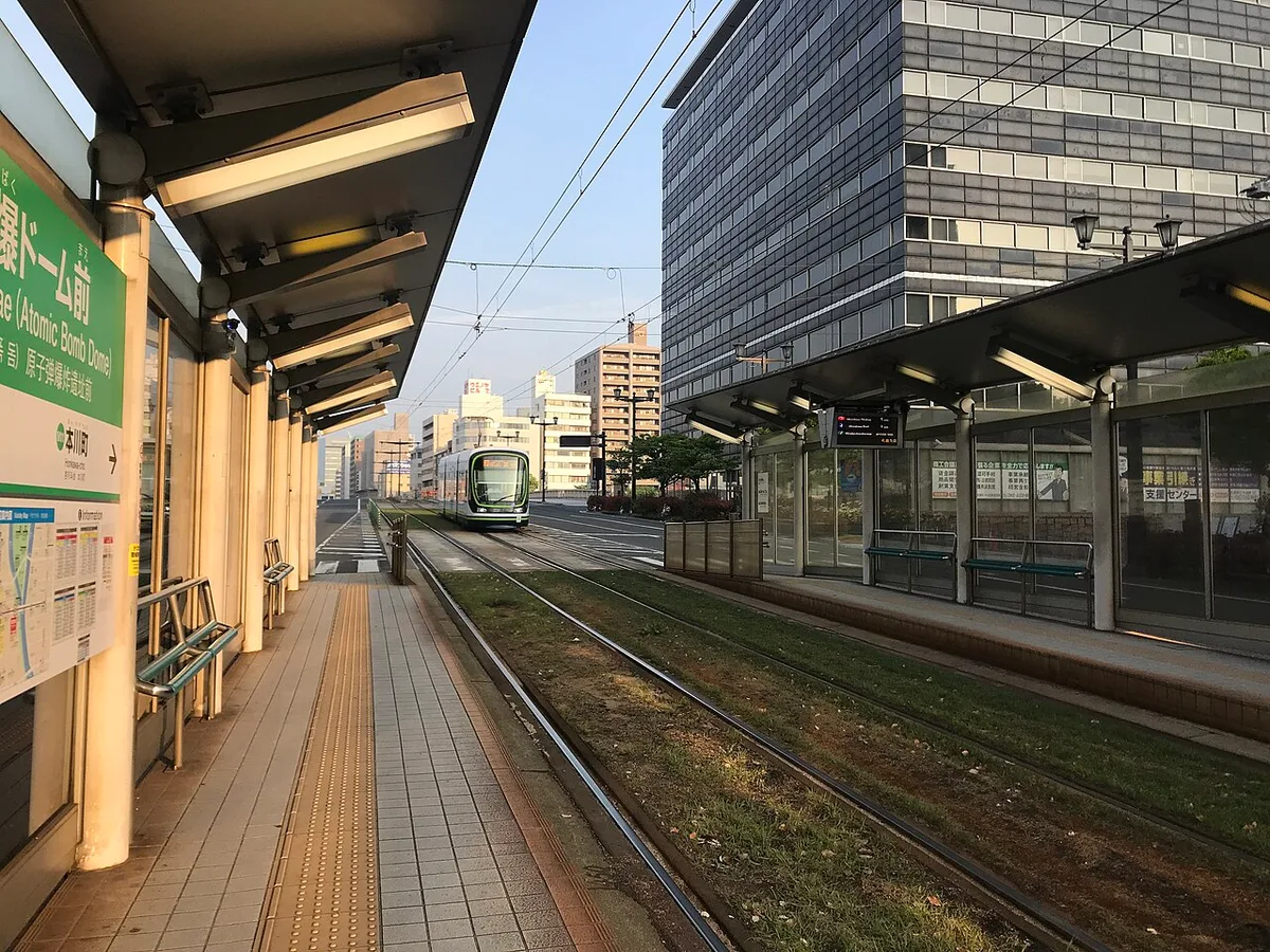 Streetcar stop with a green Genbaku Dome-mae sign and a streetcar approaching on grass-covered tracks in Hiroshima