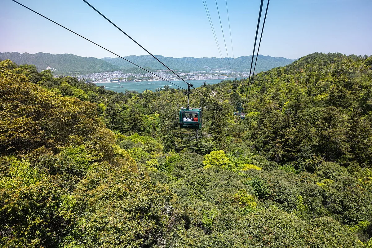 A small gondola of the Miyajima Ropeway above the forest, with the sea and the mainland coast behind