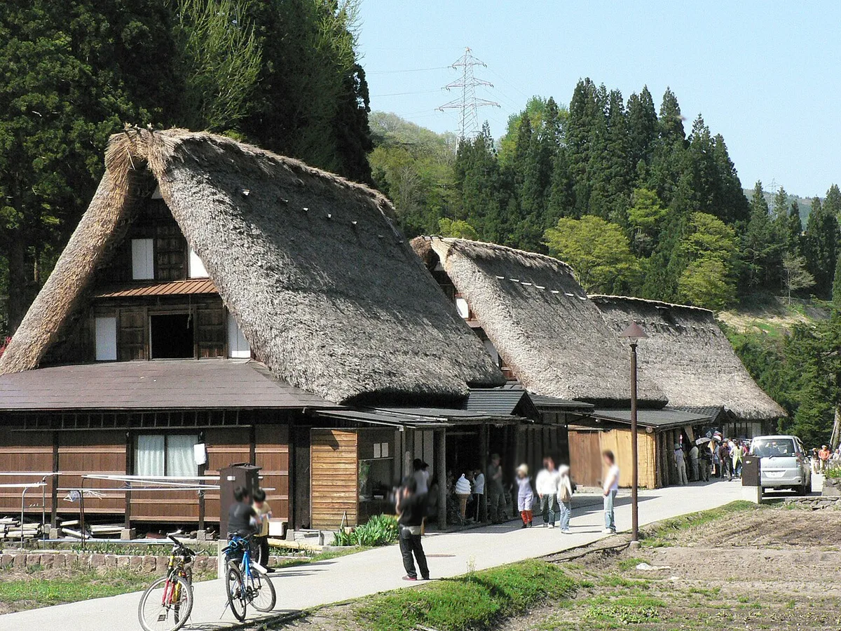 A row of thatched gassho-zukuri houses beside a paved village lane in Ainokura