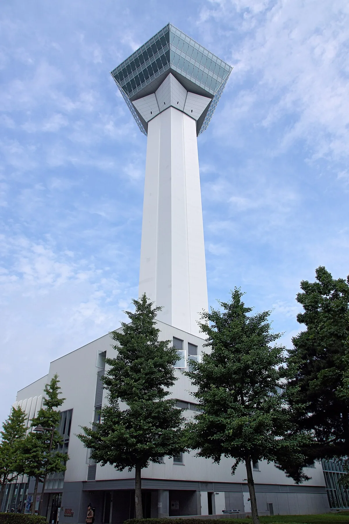 White Goryokaku Tower with its glass-walled observation deck at the top, seen from below behind a row of trees