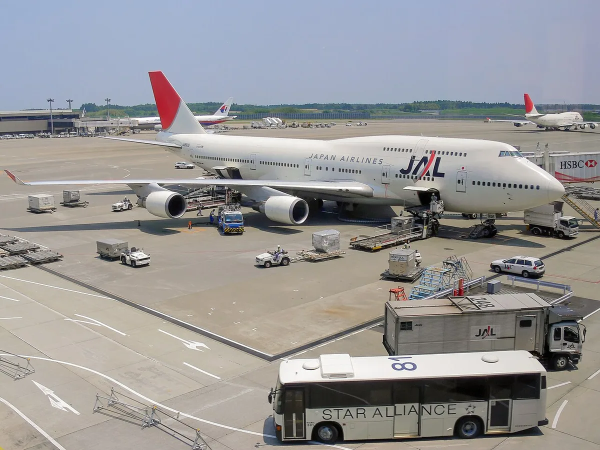 A Japan Airlines Boeing 747 parked at a gate at Narita Airport, with ground vehicles and a Star Alliance bus on the apron