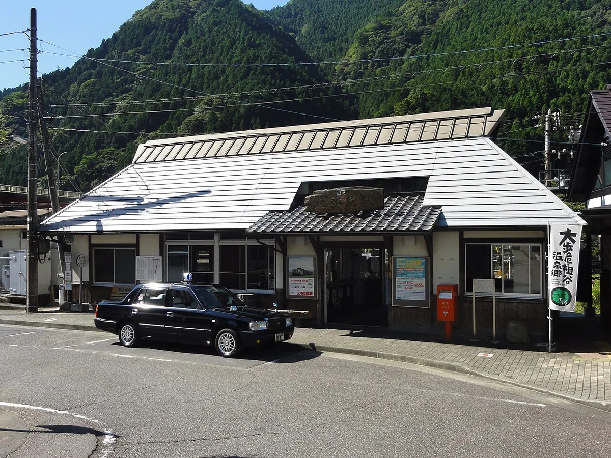 Small white-roofed JR Oboke Station building with a taxi in front and forested mountains behind
