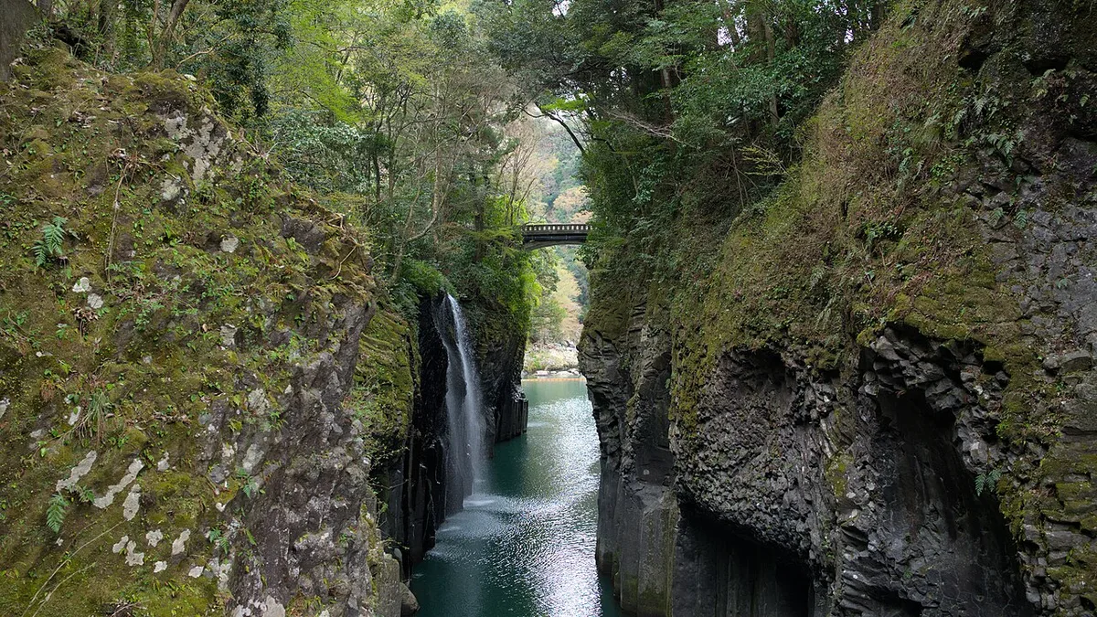 Manai Falls pouring from a mossy cliff into the green Gokase River between the basalt walls of Takachiho Gorge, with a bridge behind