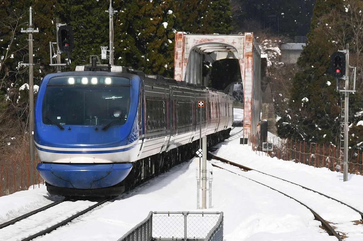 A blue and silver Super Hakuto diesel train arriving on a snowy single track by a steel bridge