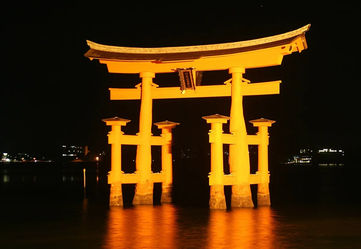 The great torii of Itsukushima Shrine lit in orange at night, standing in dark water with its reflection below