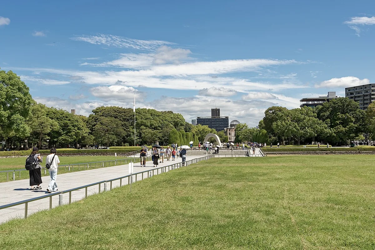 Wide lawn and paved path in Peace Memorial Park, with the arched cenotaph and the Atomic Bomb Dome in the distance under a blue sky